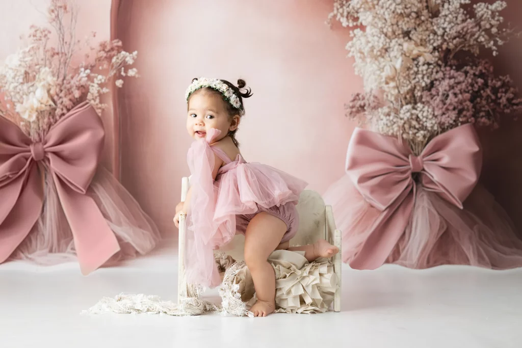 A baby wearing a pink dress and floral headband sits on a small cream bench, surrounded by large pink bows and dried floral arrangements on a pastel backdrop.