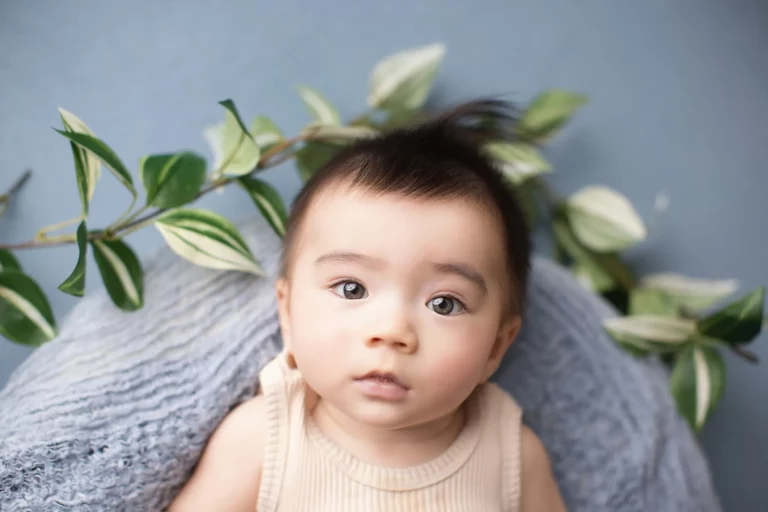 A baby with dark hair and wide eyes sits against a blue textured background with green and white variegated leaves behind them. Lovely Baby Photography, 2185 Faraday Ave suite 110, Carlsbad, CA 92008, +1 760-271-0725