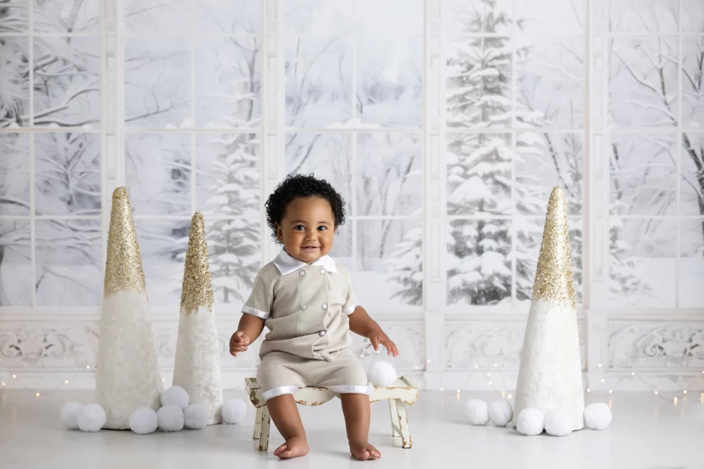 A young child in light-colored clothing sits barefoot on a small bench in front of a winter-themed backdrop with snow-covered trees and decorative white cones. Lovely Baby Photography, 2185 Faraday Ave suite 110, Carlsbad, CA 92008, +1 760-271-0725