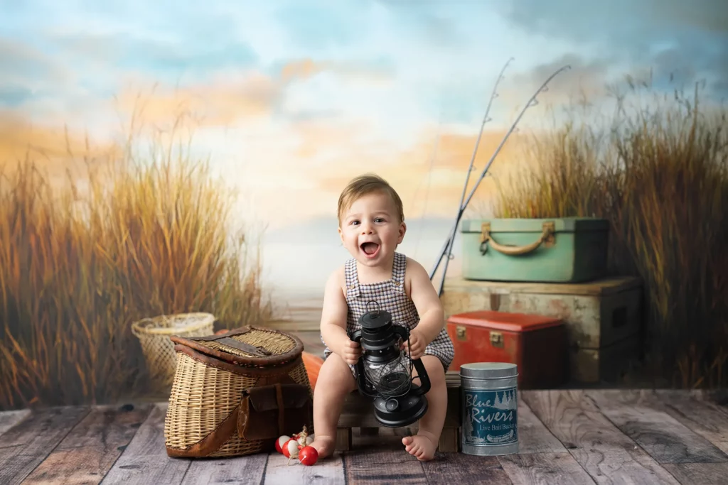 A smiling baby in overalls sits indoors with fishing props, including a lantern, baskets, tackle, and fishing rod, against a backdrop of tall grass at sunset.