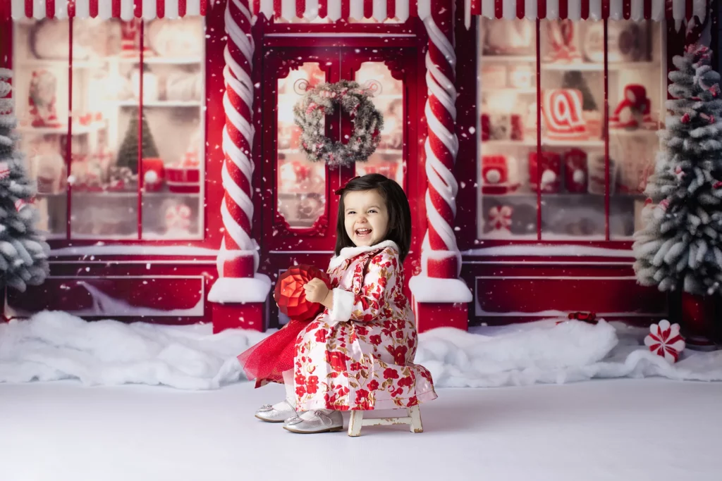 A young girl in a red and white floral dress sits on a small chair, smiling in front of a festive, winter holiday storefront backdrop with snow and Christmas decorations. Lovely Baby Photography, 2185 Faraday Ave suite 110, Carlsbad, CA 92008, +1 760-271-0725