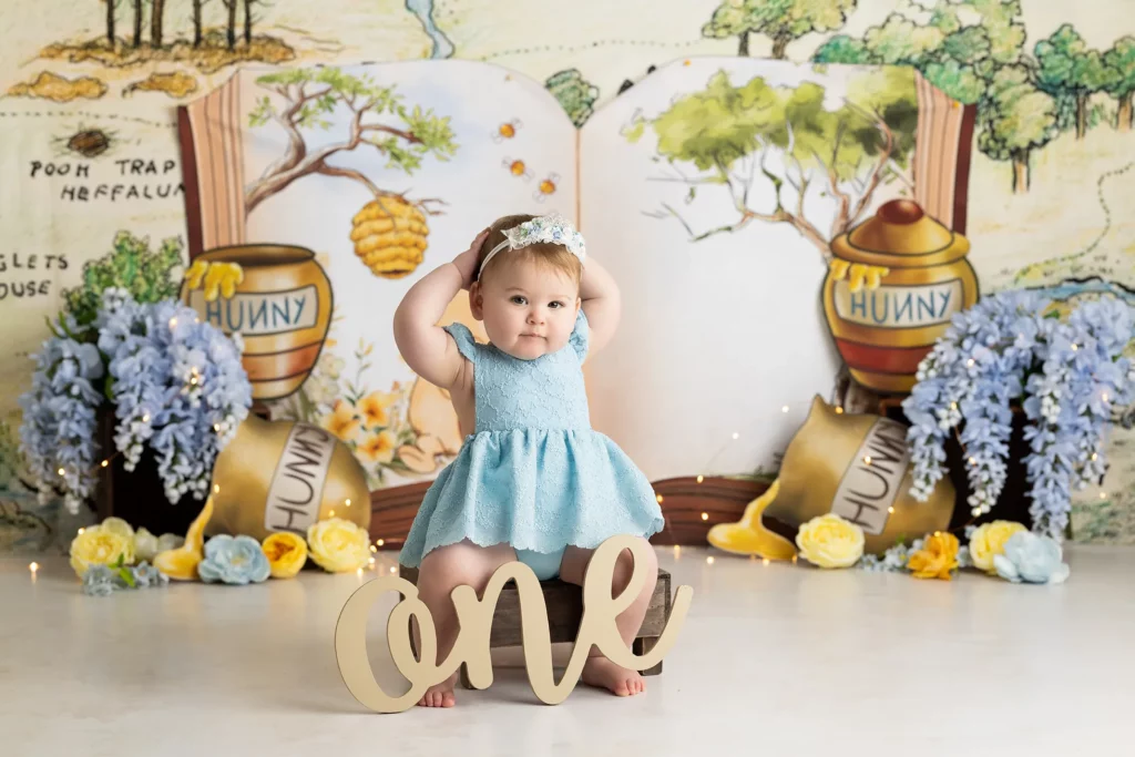 A baby in a blue dress poses with hands on head in front of a "Winnie the Pooh" themed backdrop with "Hunny" pots, flowers, and a large "one" sign.