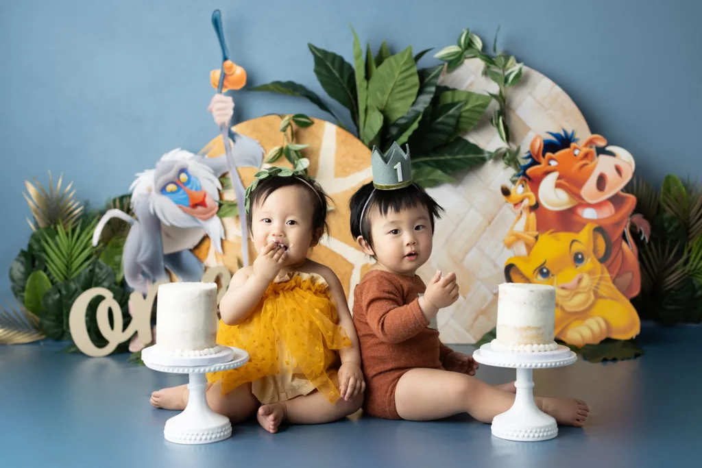 Two toddlers sit side by side in front of Lion King decorations, each with a small cake. One wears a yellow dress, the other a brown outfit and a crown with the number one.
