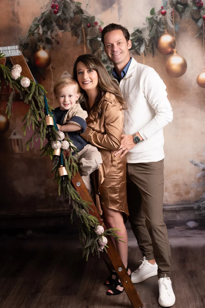 A man, woman, and young child pose together indoors beside a decorated wooden ladder with festive ornaments and greenery. Lovely Baby Photography, 2185 Faraday Ave suite 110, Carlsbad, CA 92008, +1 760-271-0725