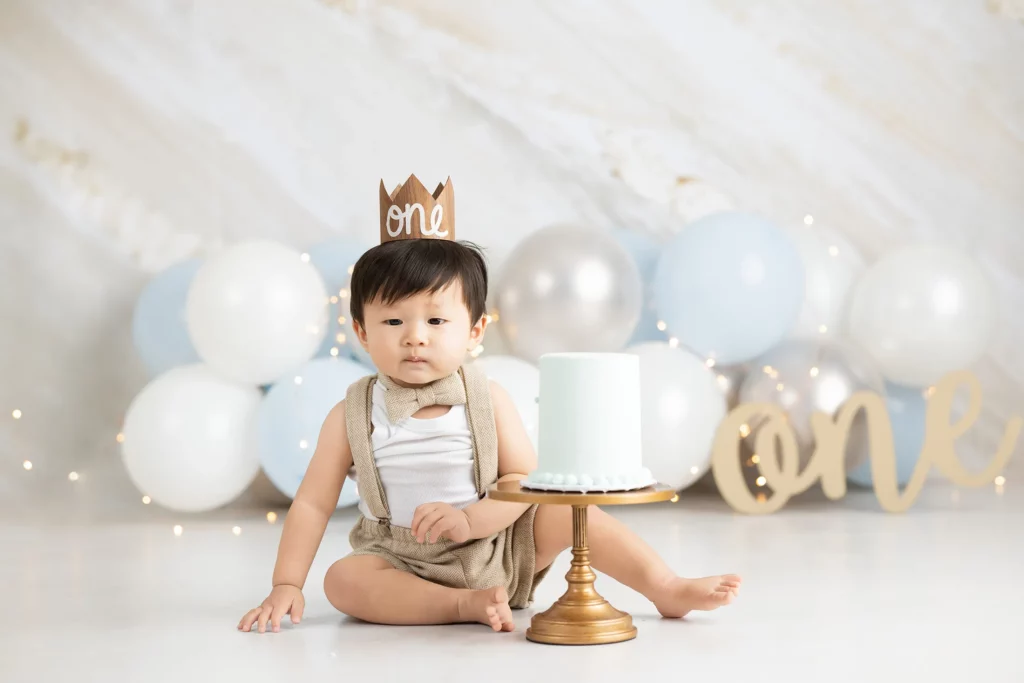 A baby wearing a crown labeled "one" sits next to a cake on a stand, with balloons and a "one" sign in the background.