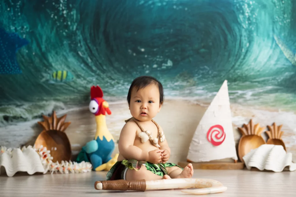 A baby dressed in a Moana-inspired costume sits on the floor with a toy paddle, surrounded by ocean-themed props, including a rooster plush and a sailboat backdrop.