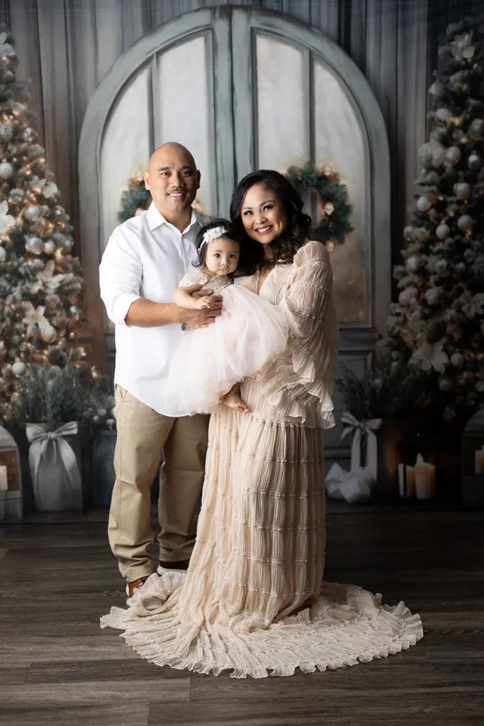 A man and woman stand together holding a baby in front of decorated Christmas trees and a large arched window backdrop, all dressed in formal light-colored clothing. Lovely Baby Photography, 2185 Faraday Ave suite 110, Carlsbad, CA 92008, +1 760-271-0725