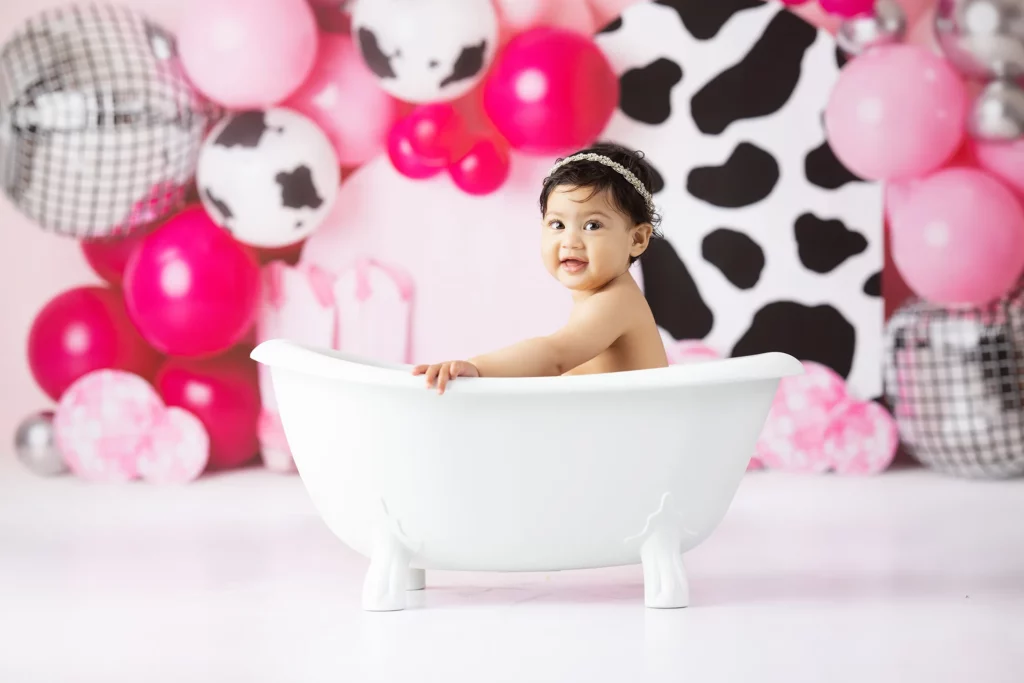 A baby sits in a white clawfoot bathtub, surrounded by pink, black, and white balloons with cow-print patterns in the background.