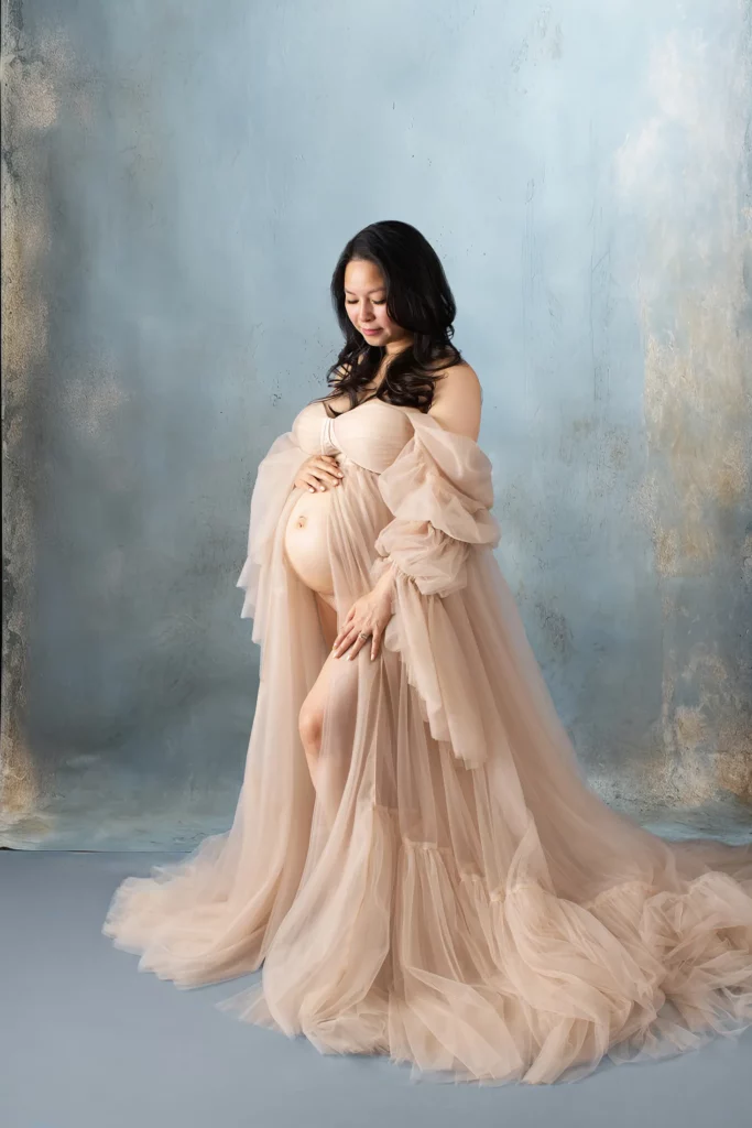 A pregnant woman in a flowing, sheer beige gown stands against a textured blue background, holding her belly and looking down with a gentle smile.