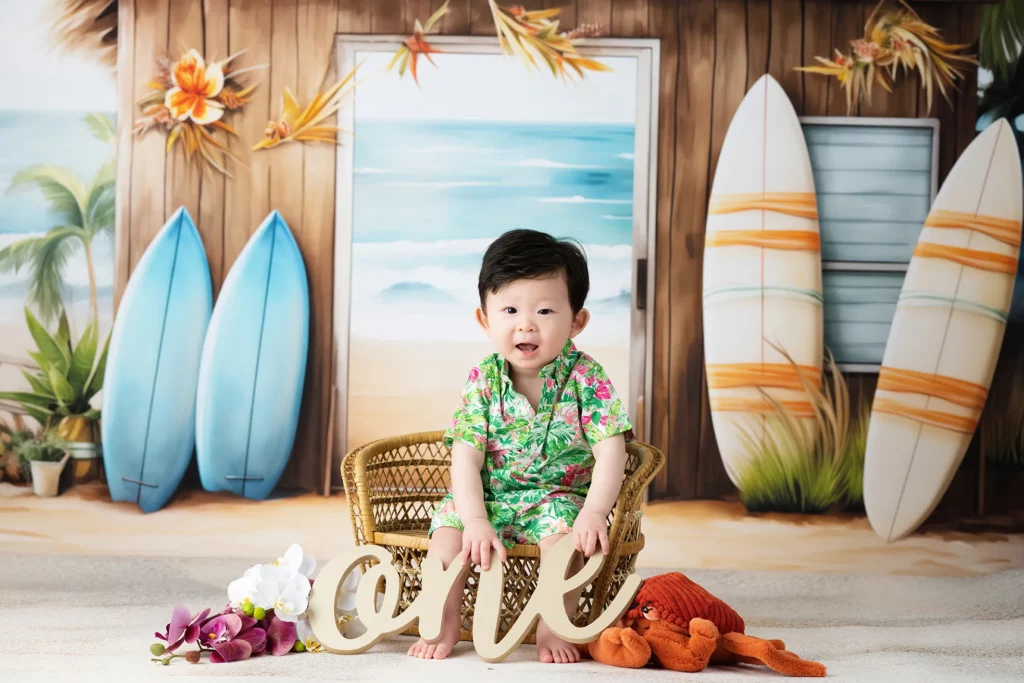 A baby in a floral outfit sits in a wicker chair holding a "one" sign, with surfboards, beach-themed decor, and flowers in the background.