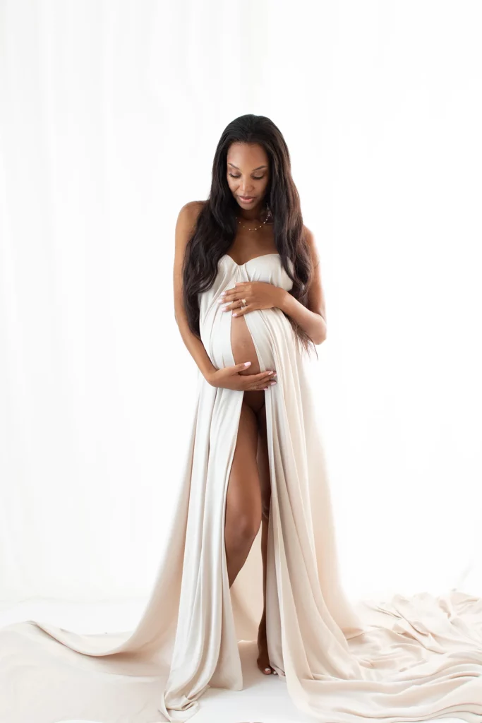 A pregnant woman in a flowing, strapless cream gown stands barefoot against a white background, gently cradling her belly and looking down.