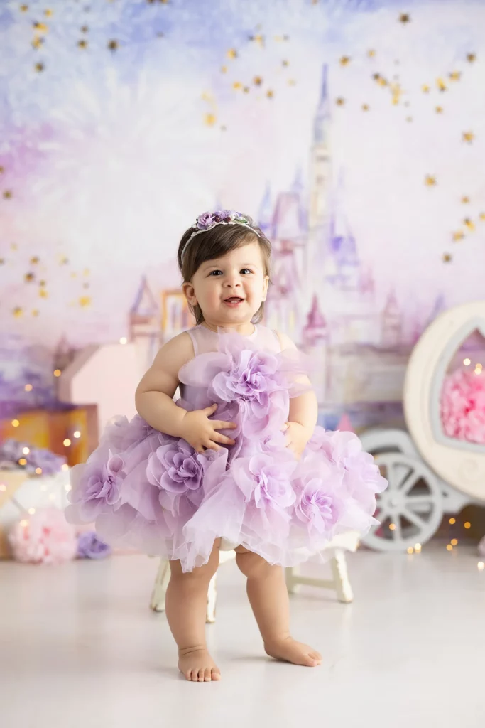 A baby wearing a lavender flower dress and headband stands barefoot in front of a fairytale castle and carriage backdrop.