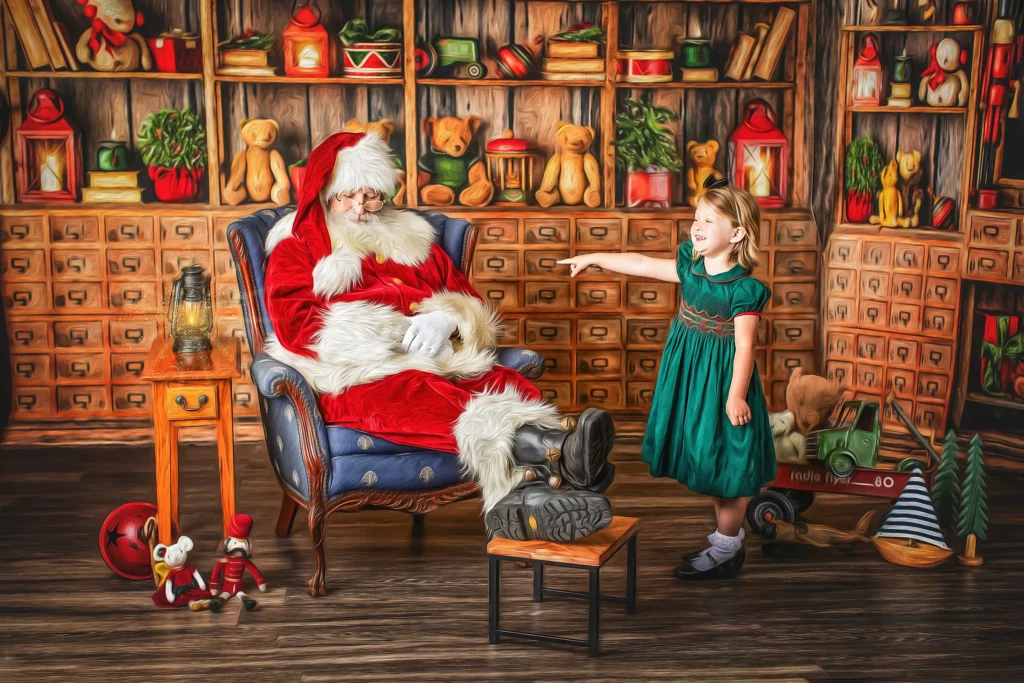 A man dressed as Santa Claus sits in a chair while a young girl in a green dress stands next to him pointing, with shelves of toys and books in the background. Lovely Baby Photography, 2185 Faraday Ave suite 110, Carlsbad, CA 92008, +1 760-271-0725