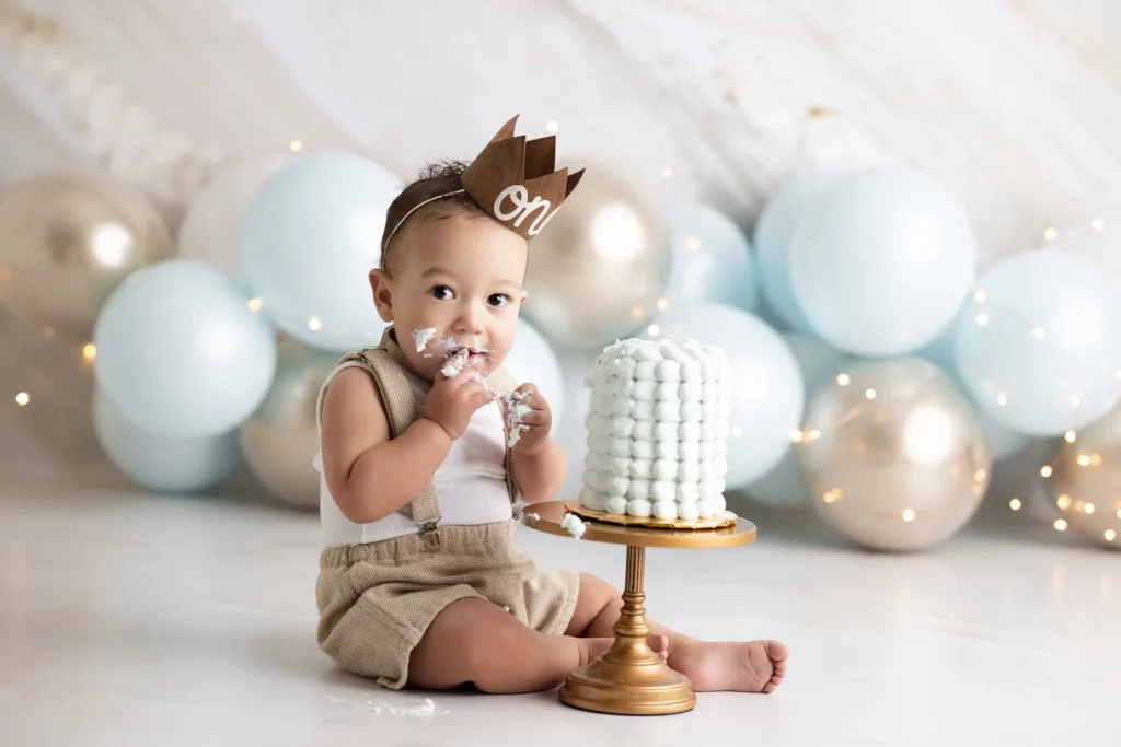 Baby wearing a "one" crown sits on the floor with a small cake, eating with hands, surrounded by blue and gold balloons. Lovely Baby Photography, 2185 Faraday Ave suite 110, Carlsbad, CA 92008, +1 760-271-0725