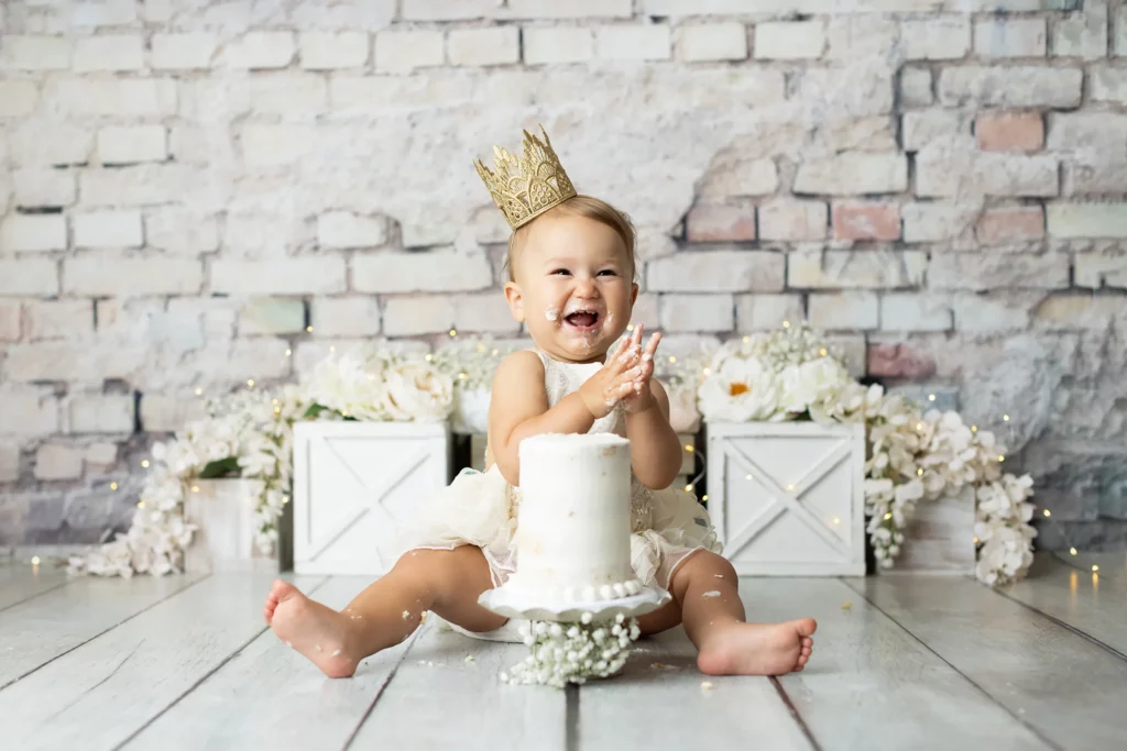 Smiling baby wearing a gold crown sits on the floor in front of a white cake, surrounded by white flowers and wooden boxes, with cake on their face and hands.