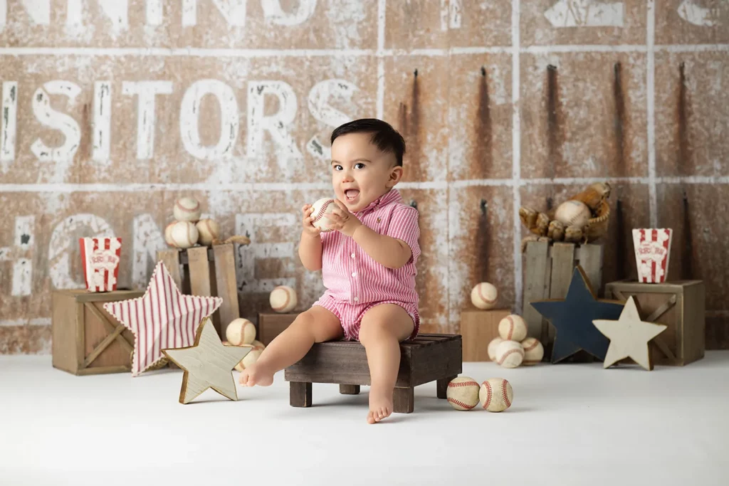 A baby in a pink striped romper sits on a small wooden bench holding a baseball, surrounded by baseballs, stars, and crates with popcorn.