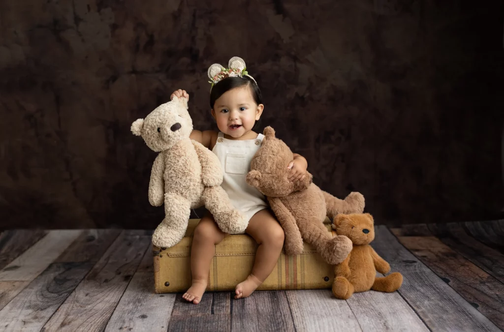 A young child wearing a floral headband sits on a suitcase, holding two teddy bears, with another teddy bear sitting beside her on a wooden floor.