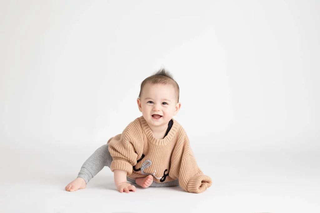 A baby wearing a tan sweater and gray pants sits on a white background, smiling and looking at the camera.