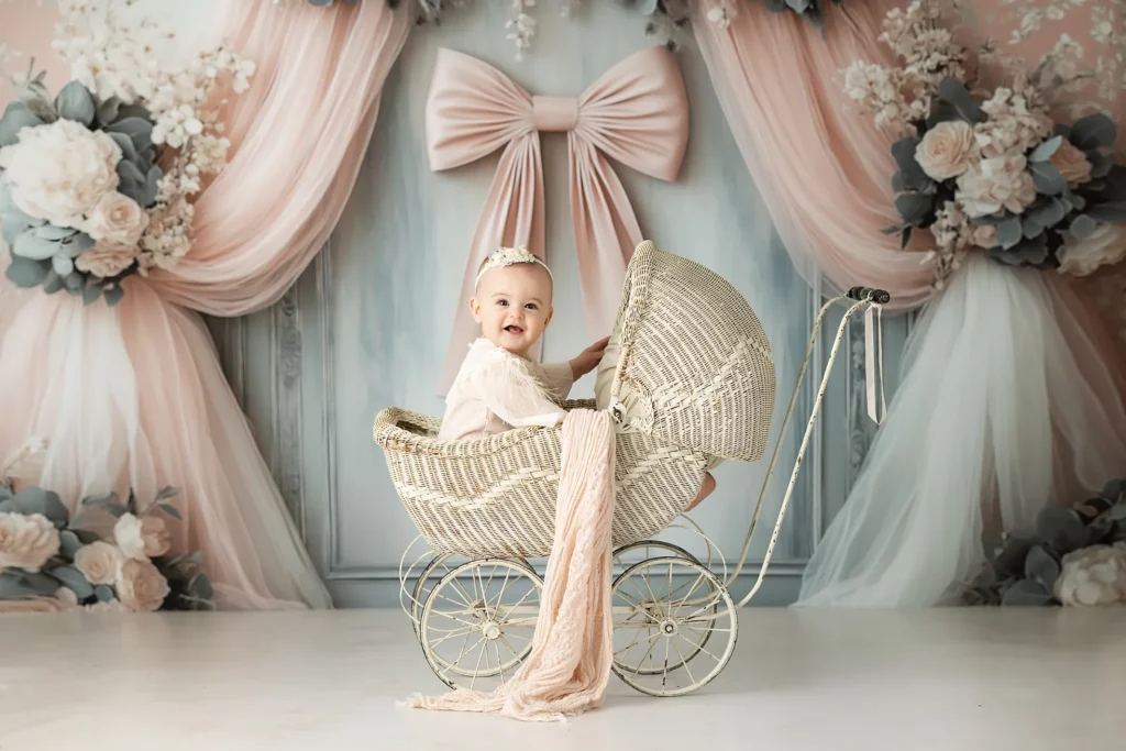 A baby sits in a vintage wicker stroller, smiling, with a soft pink sash draped over the side. The background features large pink bows and floral arrangements.