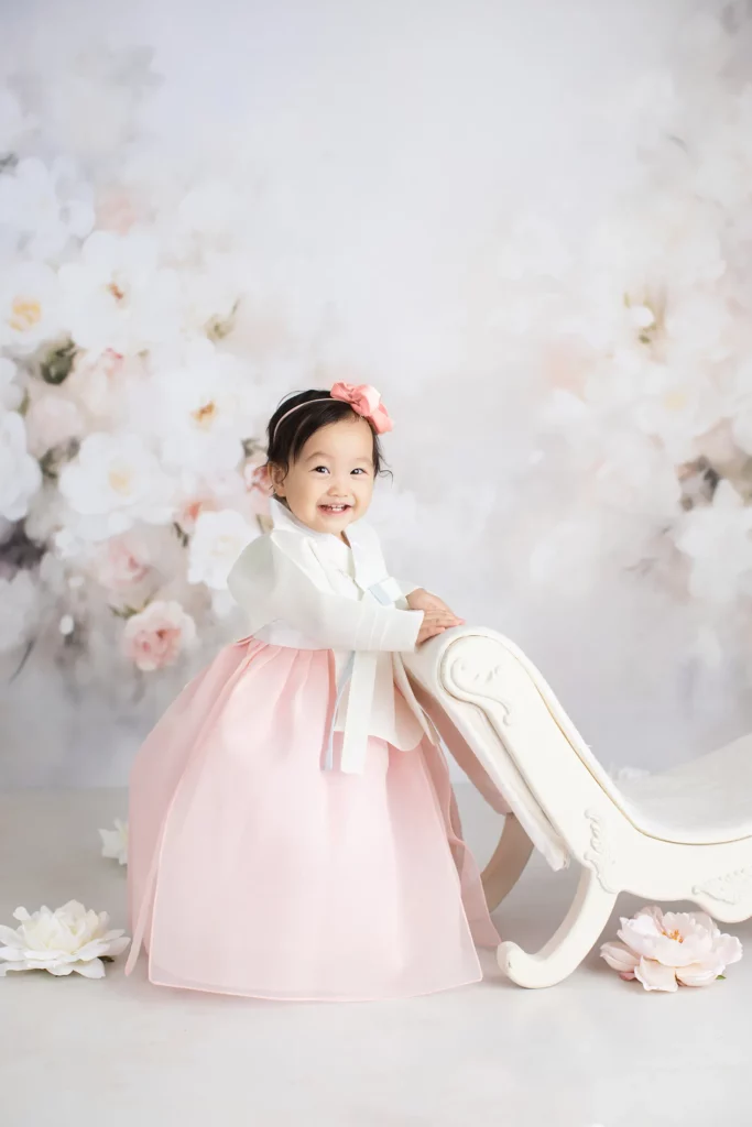 A smiling young child in a pink and white hanbok stands beside a decorative white bench, with flowers and a soft floral backdrop in the background.