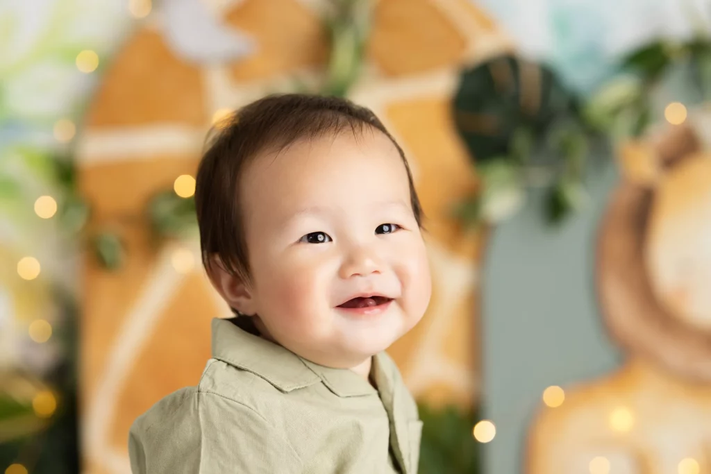 Smiling baby with short dark hair wearing a light green shirt, sitting in front of a blurred, animal-themed background with soft decorative lights.