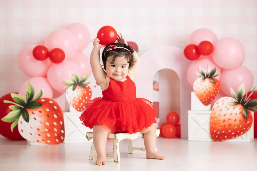 A toddler in a red dress sits on a small chair, holding a red balloon overhead. The background features pink and red balloons and large strawberry decorations.