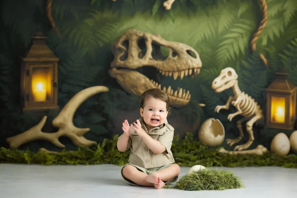 A baby sits on the floor clapping, surrounded by dinosaur skeleton props, eggs, lanterns, and greenery in a themed studio setting.