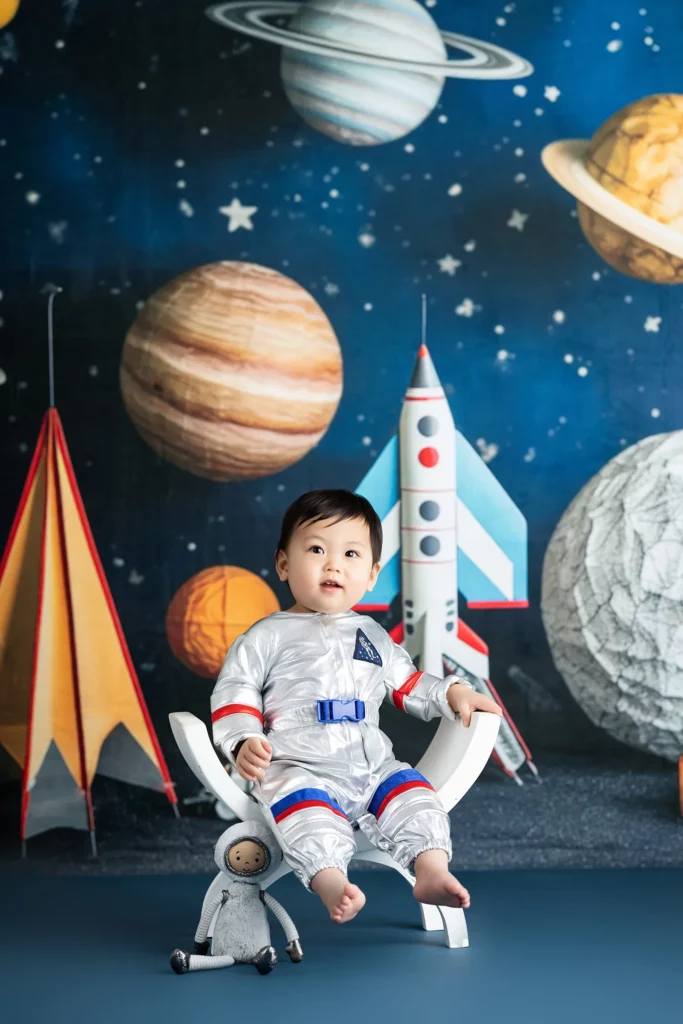 A toddler in a silver astronaut costume sits on a white chair with a stuffed toy, in front of a space-themed backdrop featuring planets, a rocket, and tents.