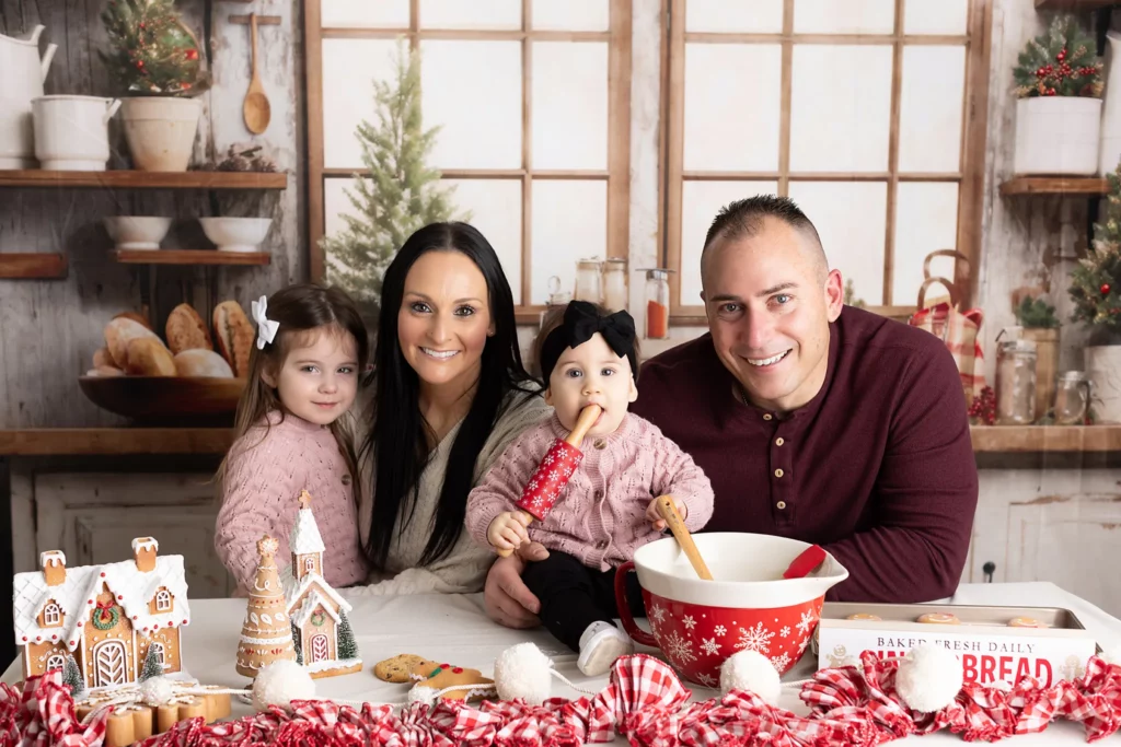 A family of four poses at a kitchen counter with holiday decorations, gingerbread houses, and baking items, smiling at the camera. Lovely Baby Photography, 2185 Faraday Ave suite 110, Carlsbad, CA 92008, +1 760-271-0725
