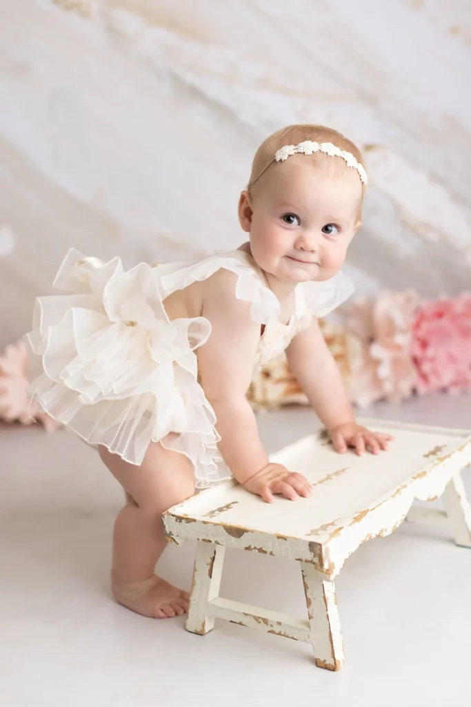 Baby wearing a white ruffled dress and headband stands holding a distressed white wooden bench with pastel flowers in the background.