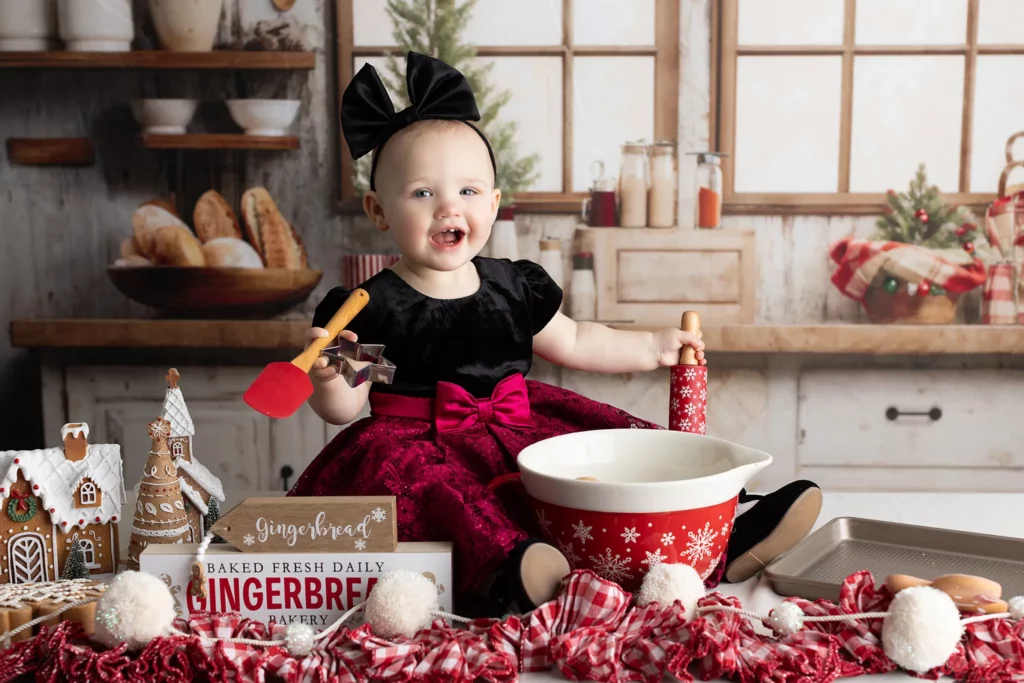 A smiling baby in a festive dress sits at a kitchen counter with baking tools, a mixing bowl, gingerbread houses, and Christmas decorations. Lovely Baby Photography, 2185 Faraday Ave suite 110, Carlsbad, CA 92008, +1 760-271-0725