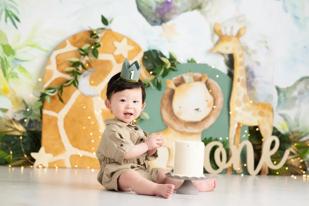 A baby wearing a crown with the number one sits next to a white cake, surrounded by jungle-themed decorations with a lion and giraffe.