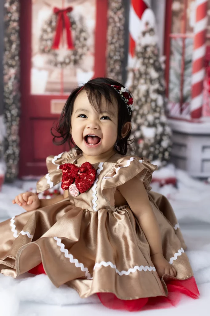 A young child in a gingerbread-themed dress with a red bow sits and smiles in front of a festive holiday backdrop with decorated trees and candy cane stripes. Lovely Baby Photography, 2185 Faraday Ave suite 110, Carlsbad, CA 92008, +1 760-271-0725