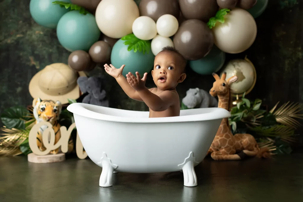 A baby sits in a white tub with arms raised, surrounded by stuffed animals and jungle-themed balloon decorations in the background.