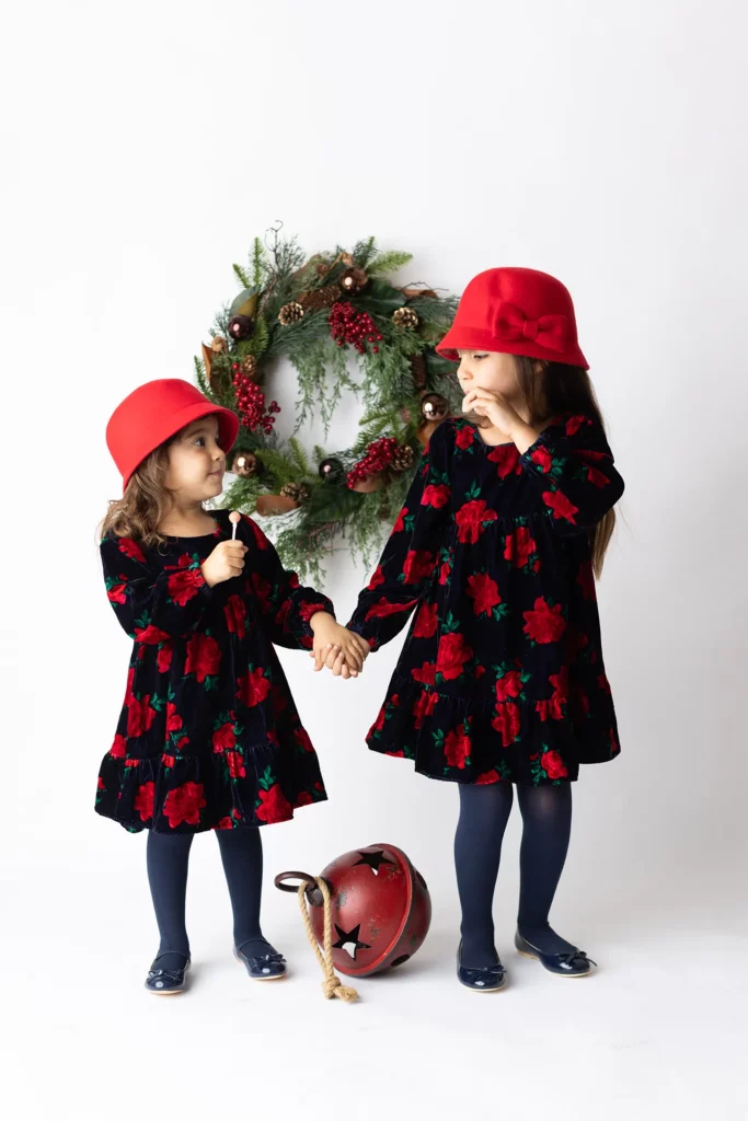 Two young girls in matching floral dresses and red hats hold hands in front of a holiday wreath, with a red sleigh bell decoration on the floor. Lovely Baby Photography, 2185 Faraday Ave suite 110, Carlsbad, CA 92008, +1 760-271-0725