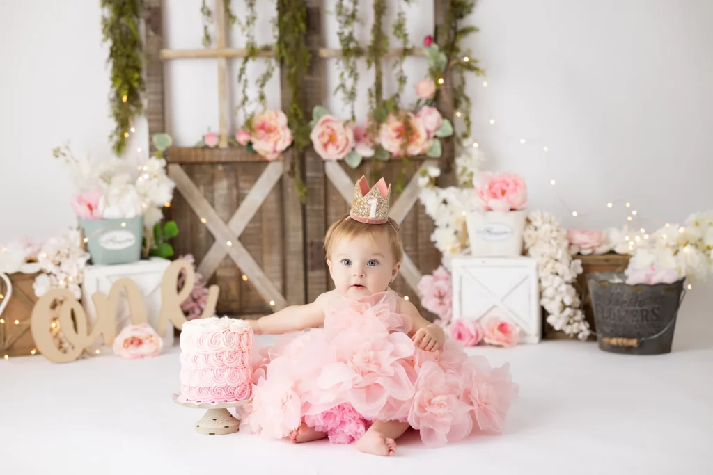 A baby in a pink dress and crown sits in front of a pink cake, surrounded by flowers, crates, and wooden decor.