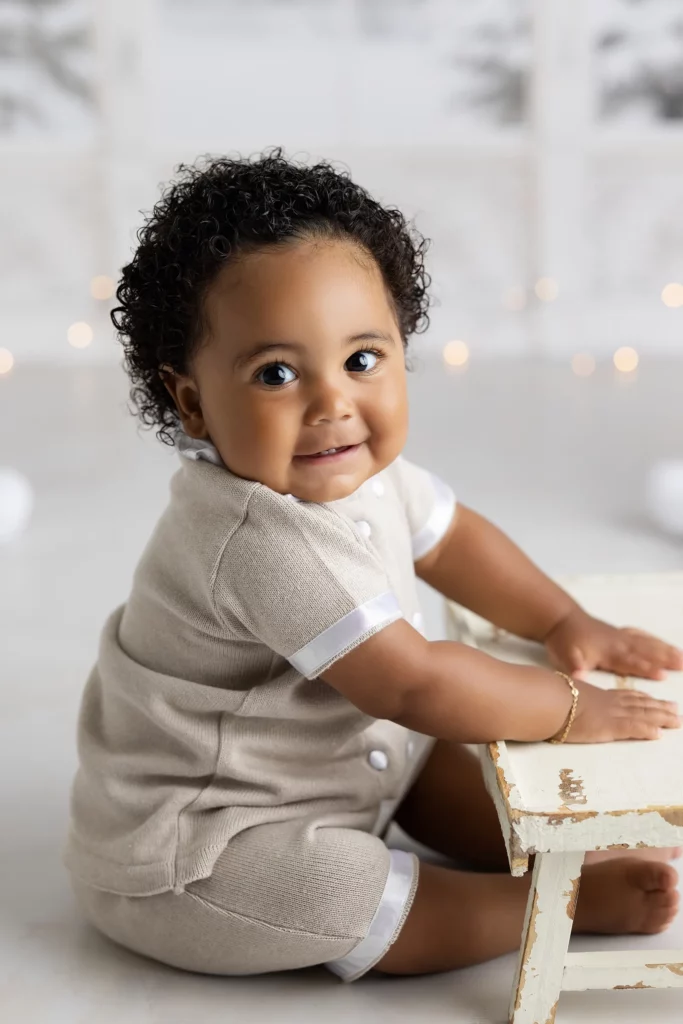Baby with curly hair in a beige outfit sits on the floor, leaning on a small white wooden bench, smiling at the camera. Lovely Baby Photography, 2185 Faraday Ave suite 110, Carlsbad, CA 92008, +1 760-271-0725