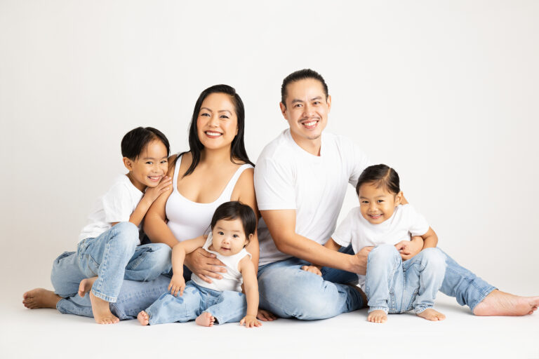 A smiling family of five poses together in a studio. The parents and three young children all wear white shirts and light blue jeans, sitting barefoot against a plain white background. Lovely Baby Photography by Stacey Marsh