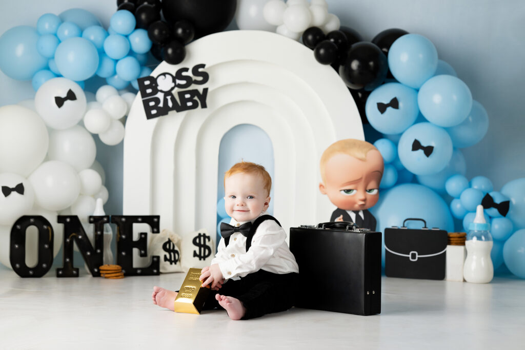 A baby in a tuxedo sits on the floor holding a gold bar prop, surrounded by blue, white, and black balloons. Behind is a "Boss Baby" sign, a "ONE" sign, and a large Boss Baby character cutout. Lovely Baby Photography by Stacey Marsh