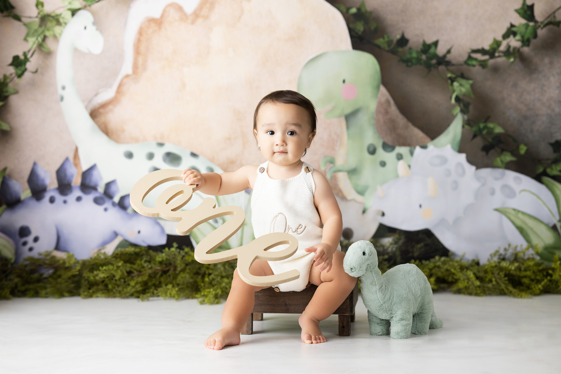 Baby boy in a white romper holding a wooden number one sign next to a dinosaur-themed first birthday backdrop in a Carlsbad studio