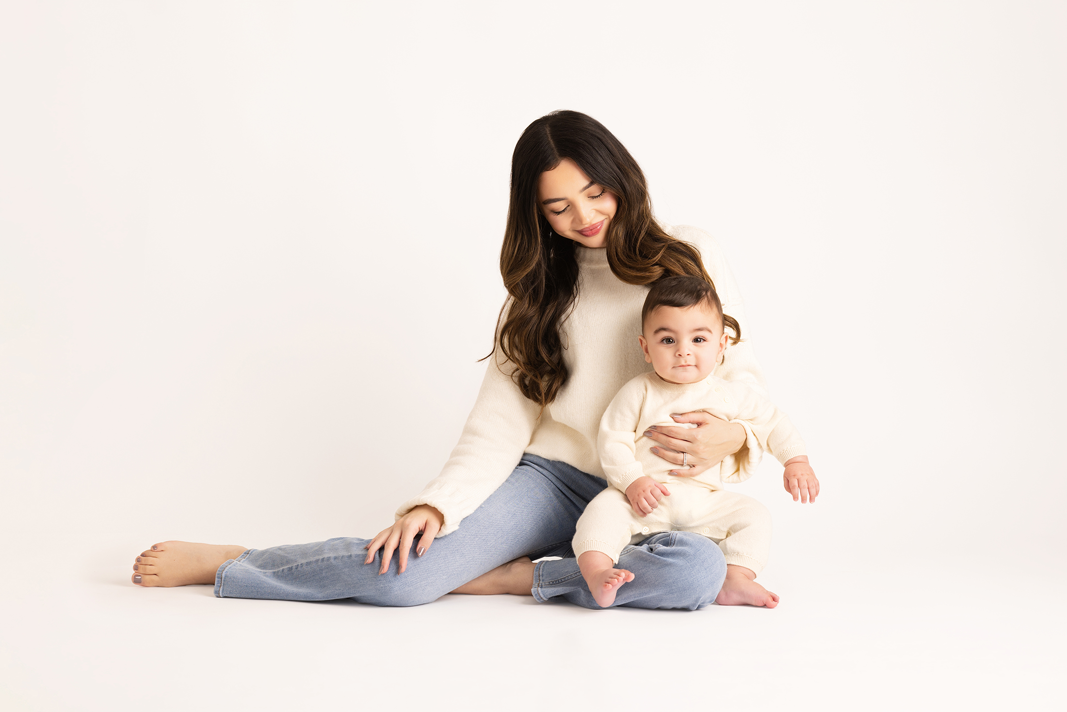A woman sits on the floor with legs crossed, holding a baby on her lap. Both wear light-colored clothes against a plain white background. Lovely Baby Photography, 2185 Faraday Ave suite 110, Carlsbad, CA 92008, +1 760-271-0725