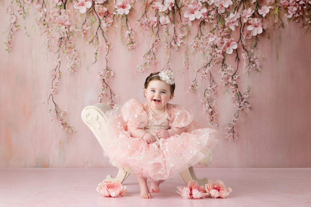 A baby girl in a pink, ruffled dress and headband sits on a white, ornate bench with pink flowers and a floral backdrop.