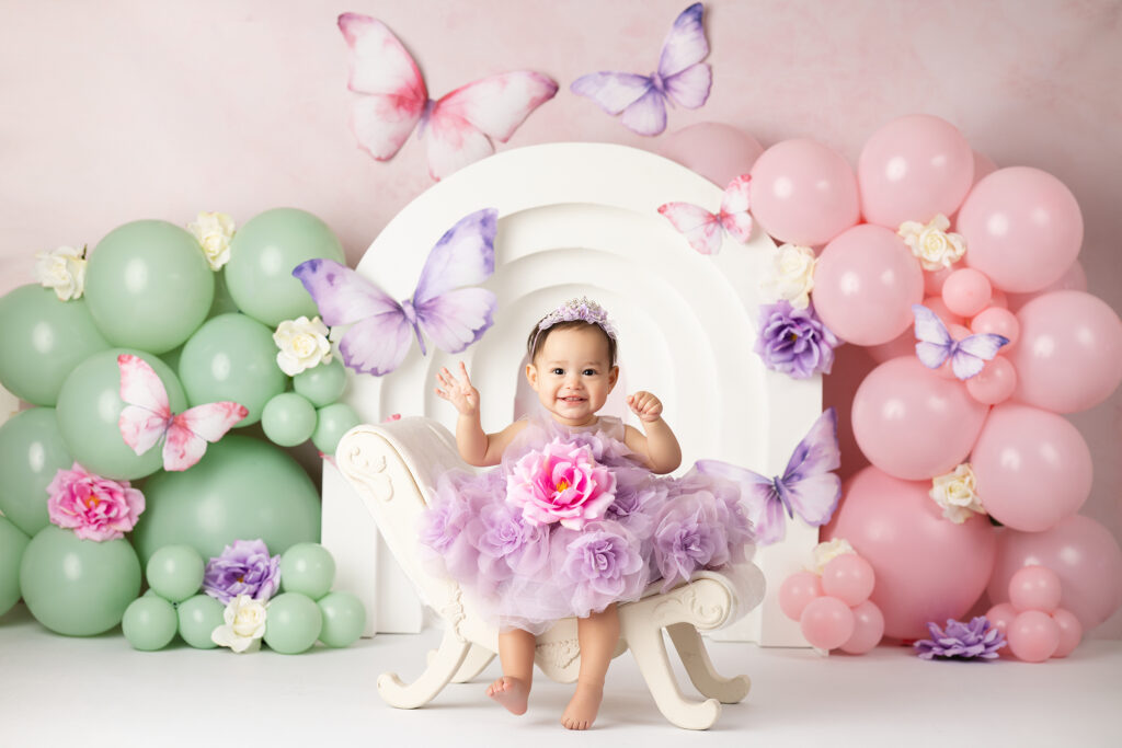 A baby in a lavender dress sits on a white chair, surrounded by pastel balloons, artificial flowers, and butterfly decorations.