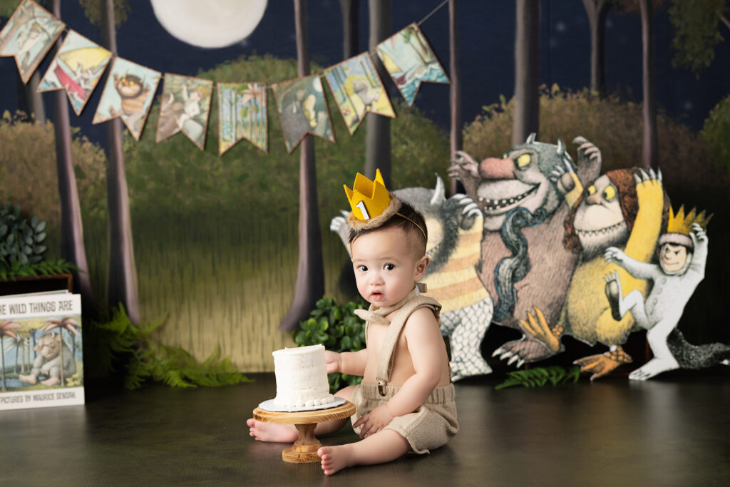 A baby wearing a crown sits on the floor next to a small cake, with "Where the Wild Things Are" themed decorations in the background.