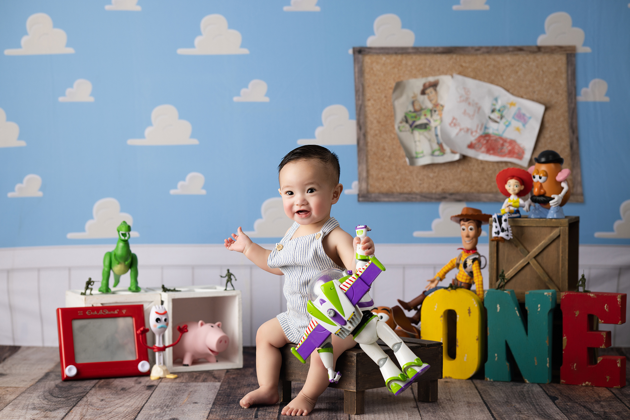 A smiling baby sits on a wooden box labeled "ONE," surrounded by Toy Story characters and toys against a blue sky and clouds backdrop. Lovely Baby Photography, 2185 Faraday Ave suite 110, Carlsbad, CA 92008, +1 760-271-0725