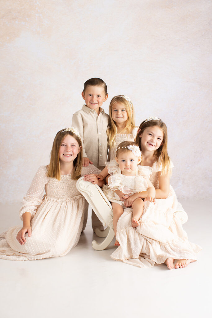 Five young children in light-colored, formal outfits pose together in a studio, seated and standing around a white bench against a neutral background. Lovely Baby Photography, 2185 Faraday Ave suite 110, Carlsbad, CA 92008, +1 760-271-0725