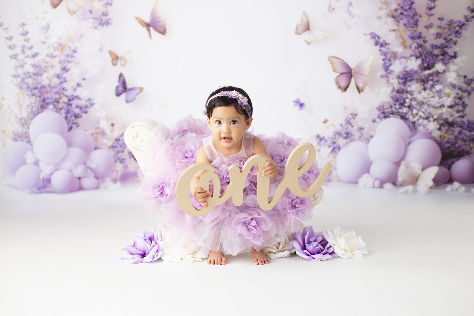 A baby in a purple tutu dress sits on a white chair holding a sign that says "one," surrounded by purple flowers, balloons, and butterfly decorations—perfectly capturing the magic of this milestone thanks to a talented first birthday photographer Carlsbad.