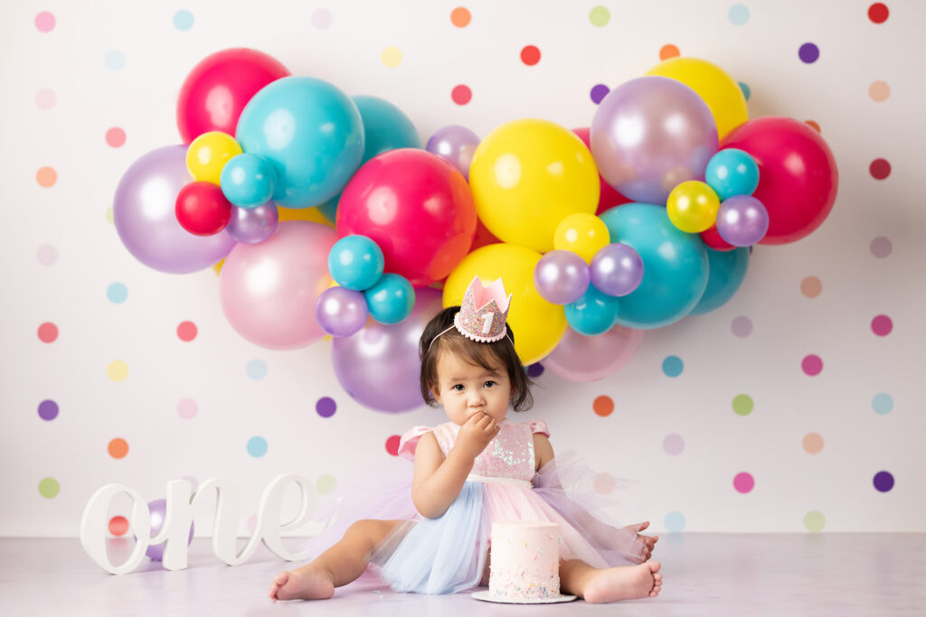 Toddler in a pastel dress and crown sits on the floor with a cake, colorful balloon garland, and "one" sign; polka dot wall background—perfectly captured by a first birthday photographer Carlsbad. Lovely Baby Photography, 2185 Faraday Ave suite 110, Carlsbad, CA 92008, +1 760-271-0725