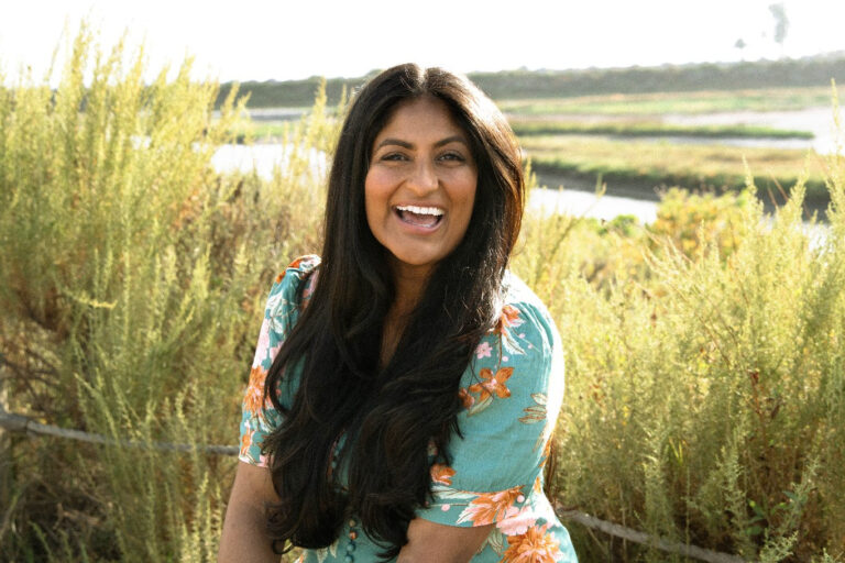 Woman with long dark hair in a floral dress smiling outdoors, standing in front of tall green plants with a blurred landscape in the background. Lovely Baby Photography, 2185 Faraday Ave suite 110, Carlsbad, CA 92008, +1 760-271-0725