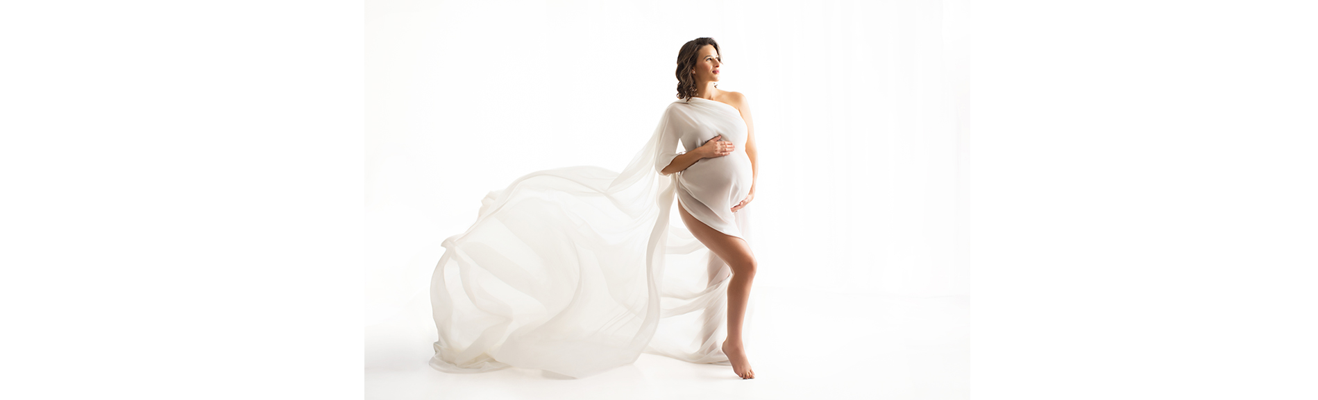 Pregnant woman standing barefoot, draped in flowing white fabric, looking to the side against a white background.